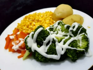 Plate of steamed broccoli, corn, carrots, and potatoes topped with yogurt dressing — a fiber-rich vegetarian meal that supports digestive health.
